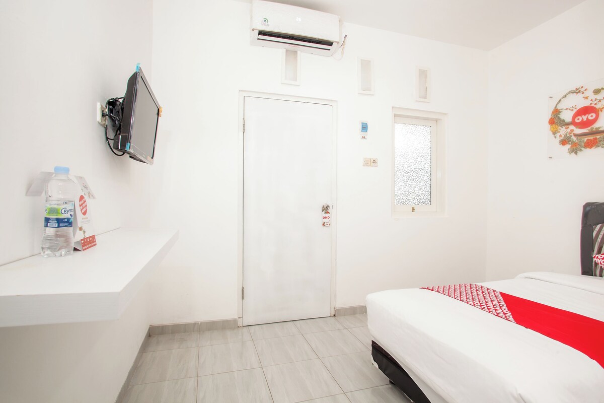 A hotel room with a bed, a white wall, a door, an air conditioning unit, a small window, and a water bottle on a shelf.