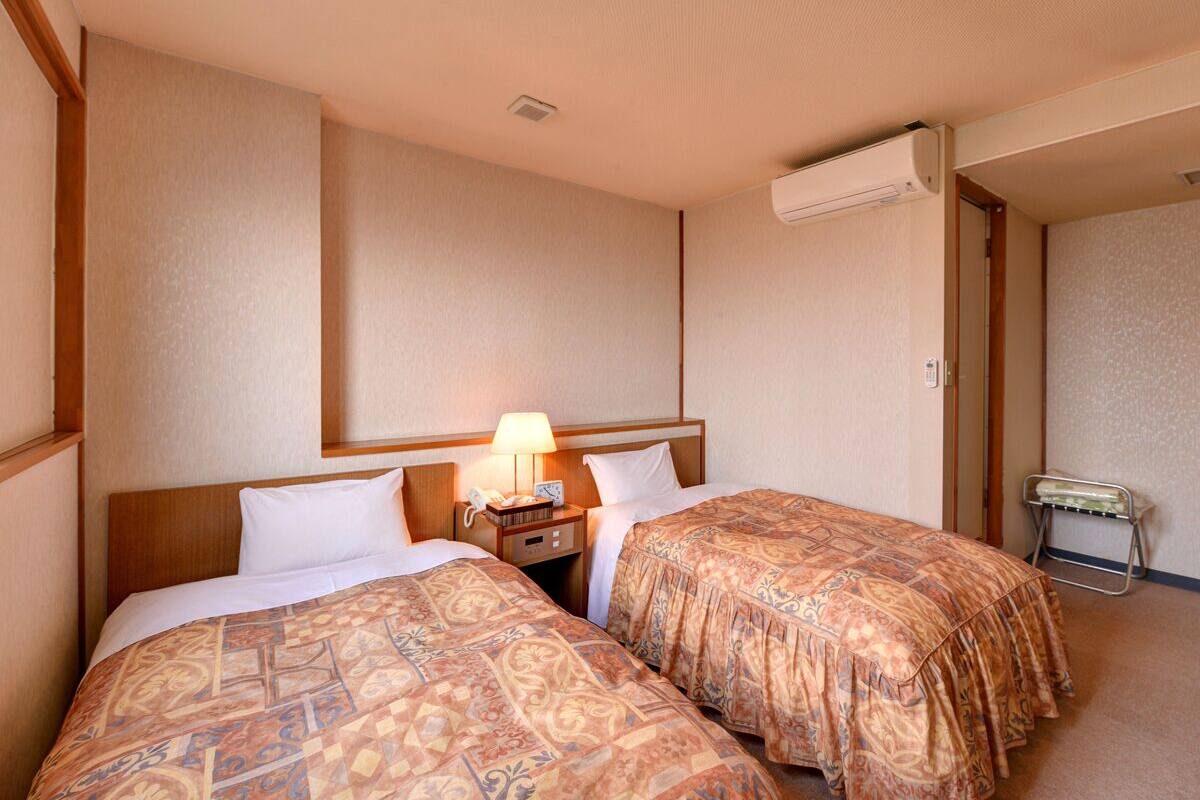 A hotel room with two beds, a nightstand with a lamp, and an air conditioning unit on the wall.