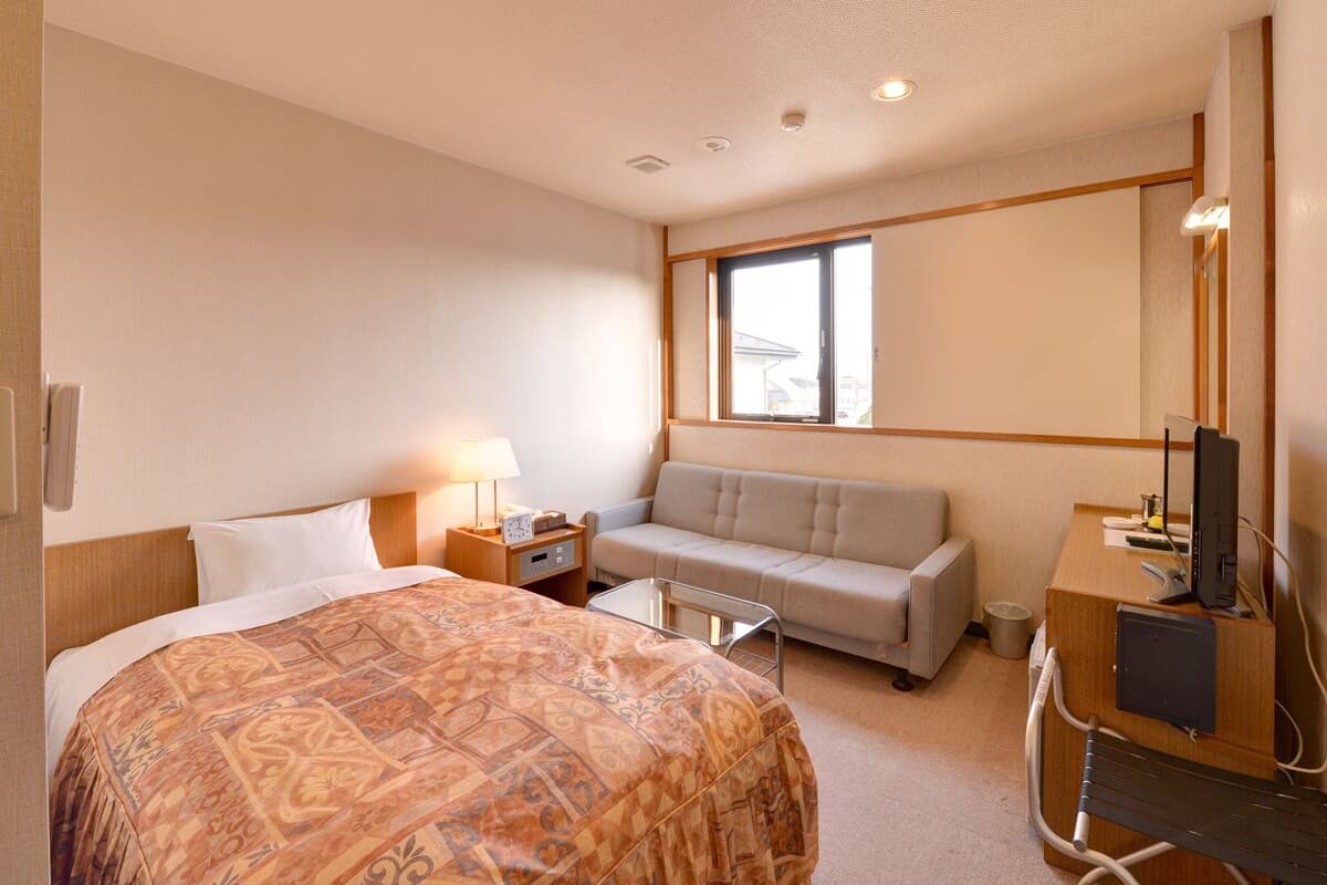 A hotel room with a large bed, a sofa, a desk with a computer, and a window providing natural light.