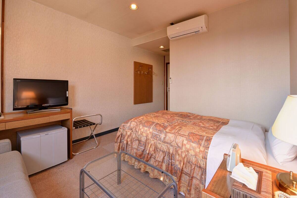 A hotel room with a bed, a desk with a television, a mini-fridge, a chair, and an air conditioning unit.
