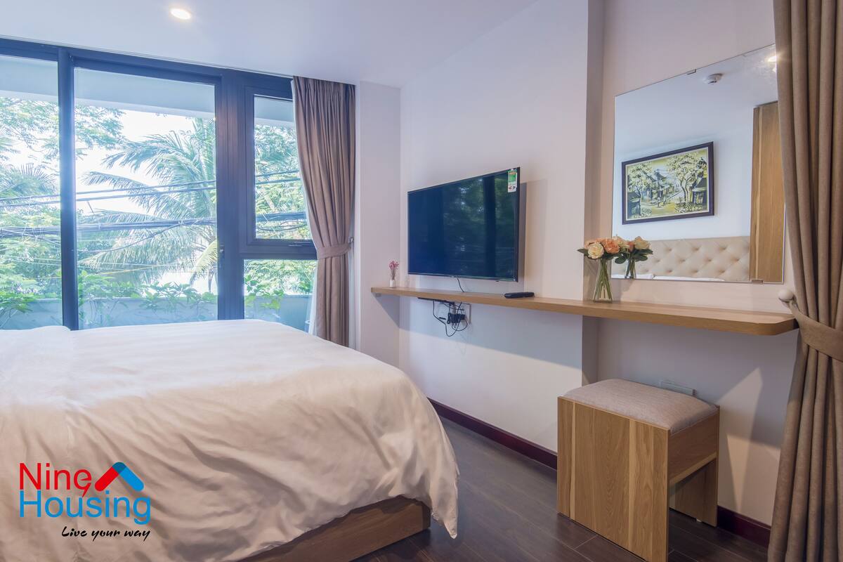 A modern hotel room with a large bed, a TV mounted on the wall, a small bench, and a window with curtains.