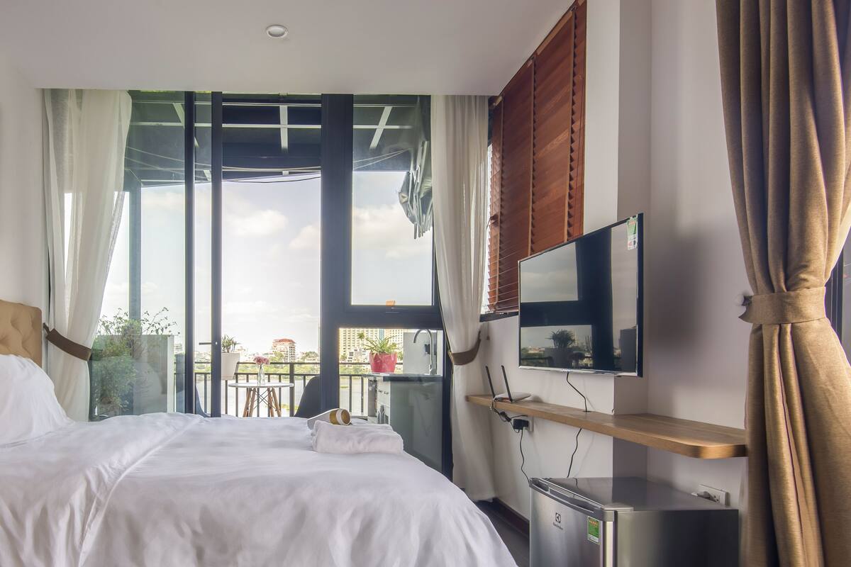 A modern hotel room with a large bed, a desk with a computer, a TV, and a balcony with a view of the city.