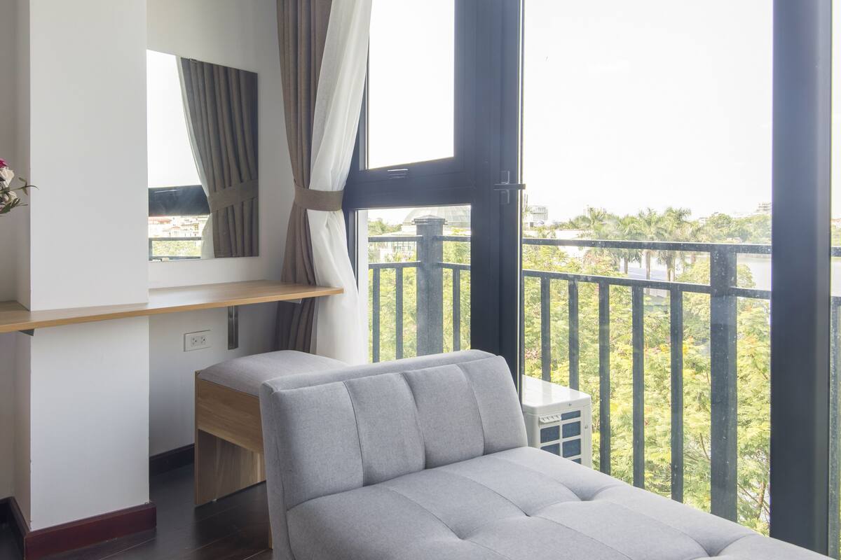 A modern bedroom with a grey sofa bed, a wooden shelf, and a large window with curtains.