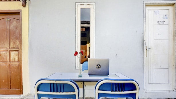Una pequeña mesa con una computadora portátil, un florero con flores, dos sillas azules y un espejo en la pared.