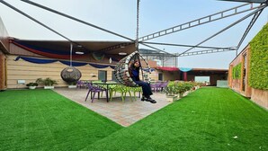 Una persona sentada en una silla colgante en un patio con césped artificial, rodeada de mobiliario de exterior y plantas en macetas.