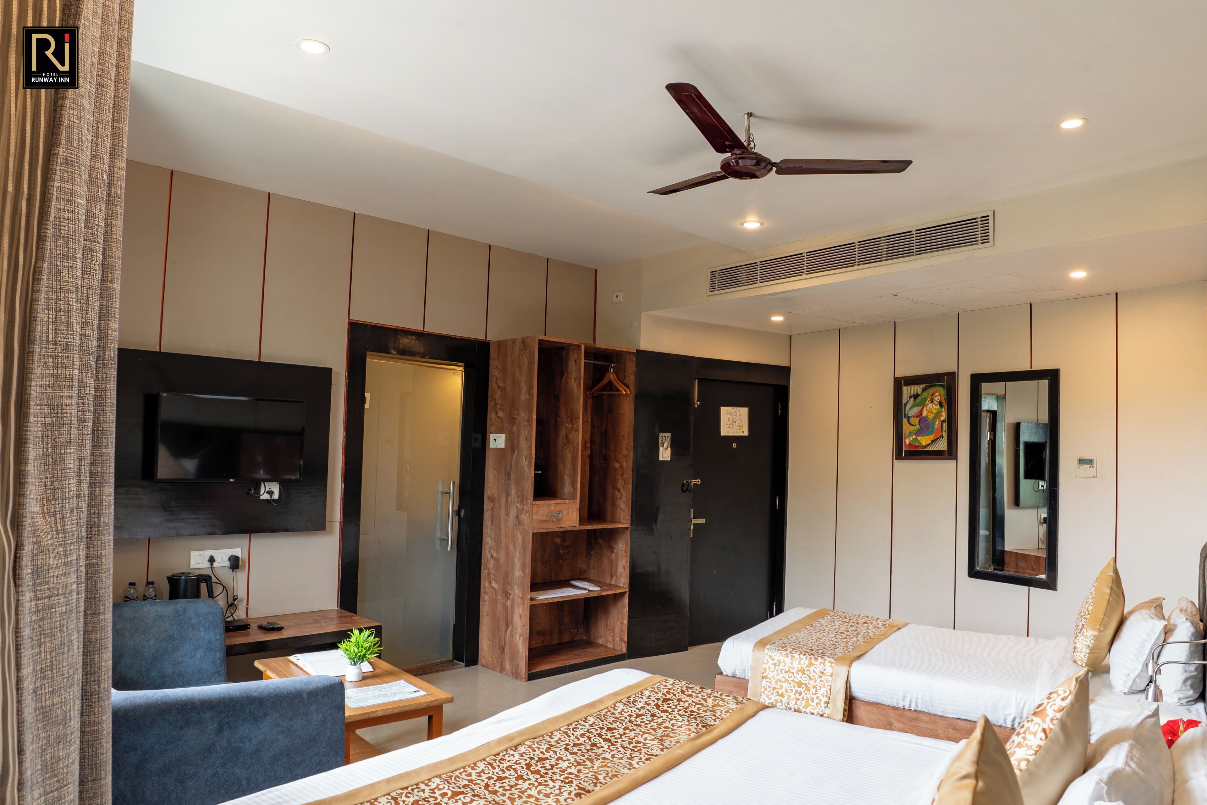A hotel room with a flat-screen TV, a ceiling fan, a wardrobe, a desk with a chair, and two beds with decorative bedding.