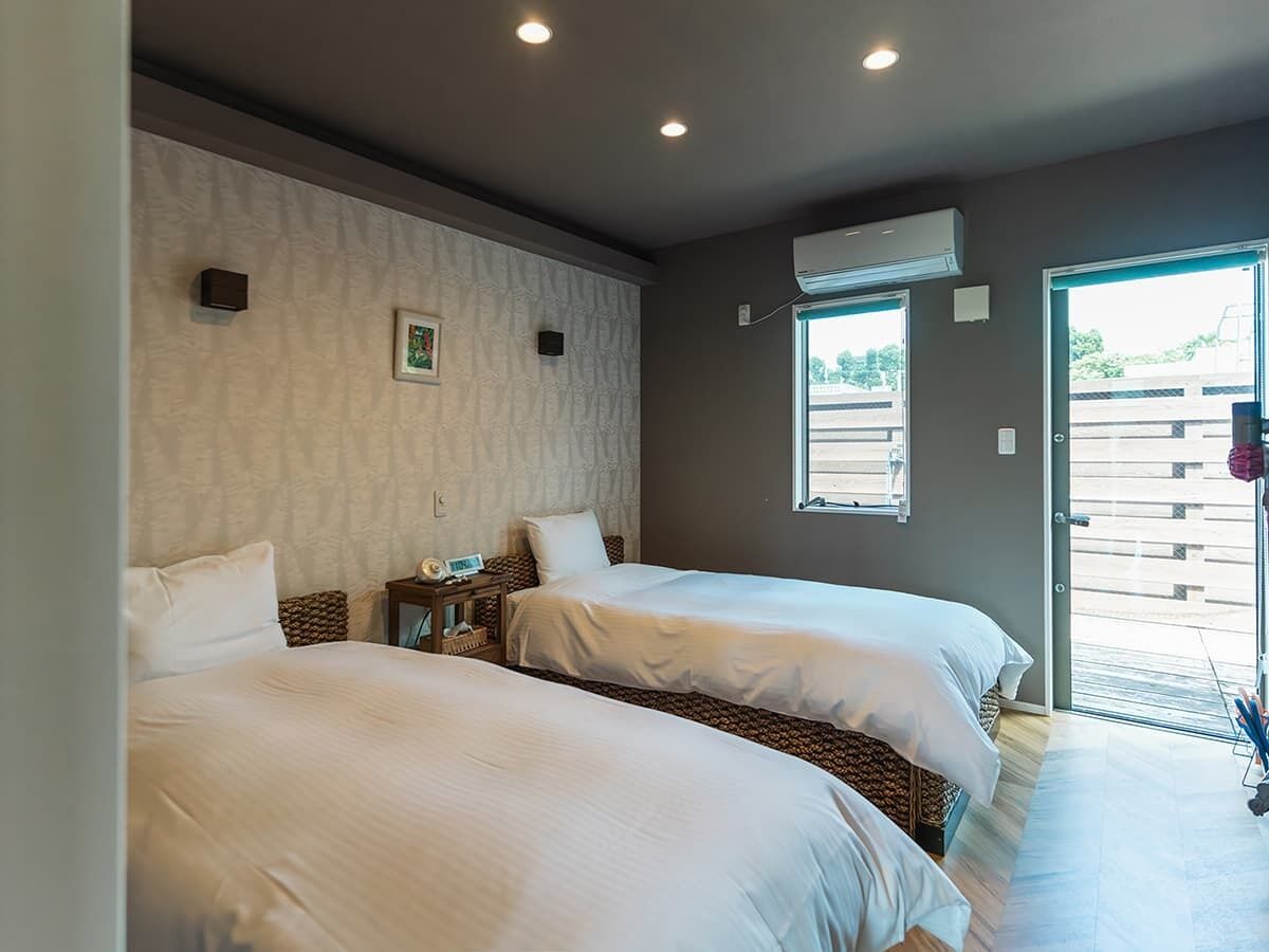 Ein Hotelzimmer mit zwei Betten, einem kleinen Nachttisch mit Lampe, einer Klimaanlage und einem Fenster mit Blick nach draußen.