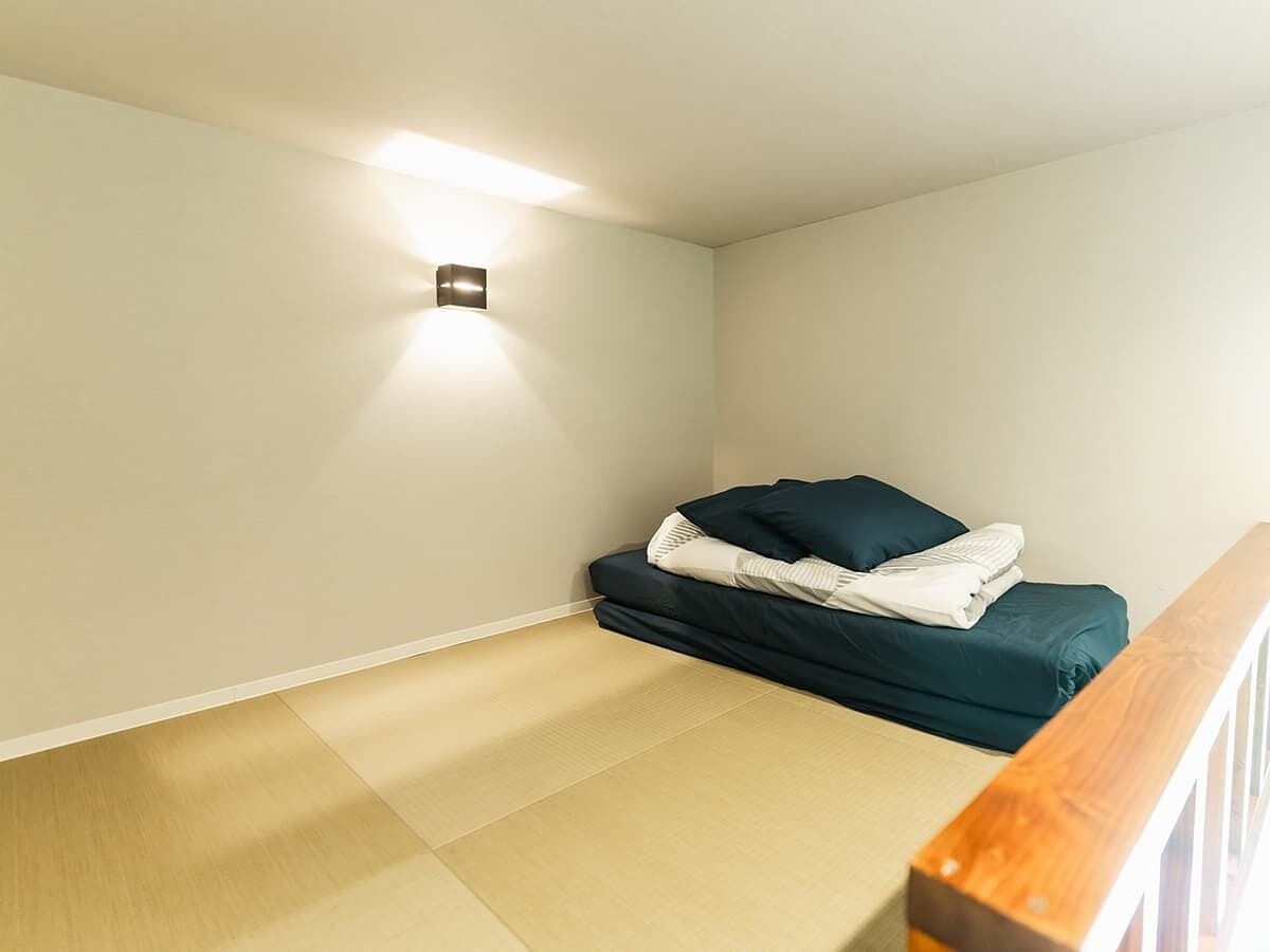 Une pièce avec un sol recouvert de tatamis, un lit bas surmonté d’un matelas vert foncé et un petit luminaire fixé au mur.