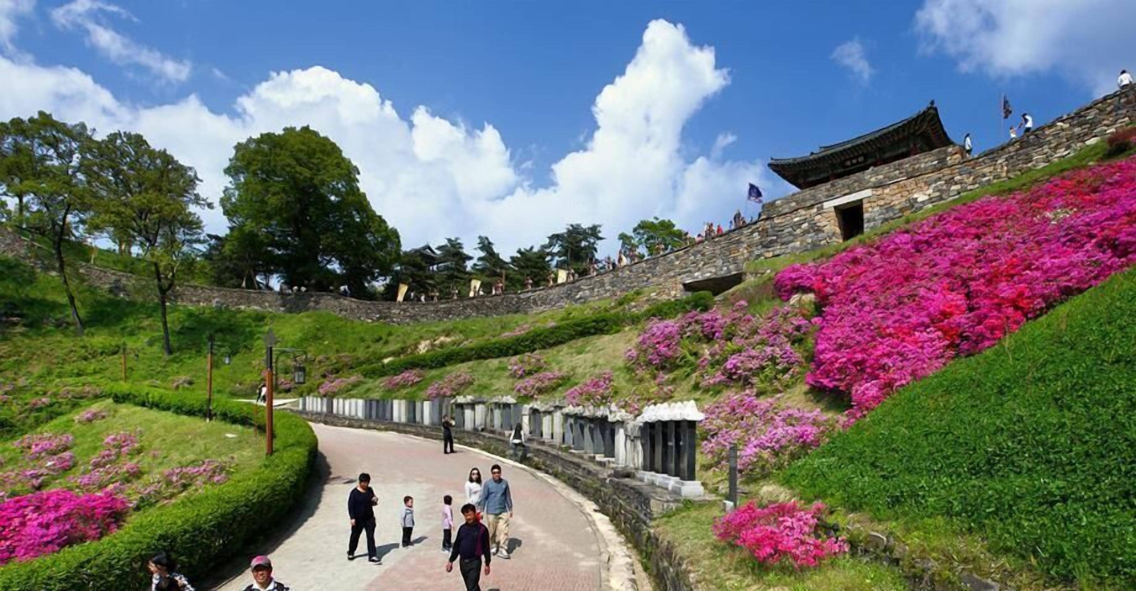 A pathway with people walking, surrounded by greenery and pink flowers, leading to a traditional building with a tiled roof.