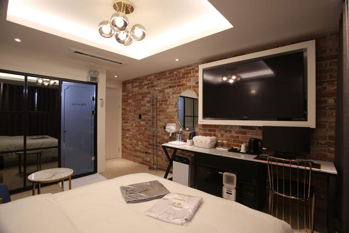 Quarto de hotel moderno com uma cama grande, uma mesa com computador, TV e parede de tijolos.