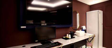 A modern hotel room with a large flat-screen TV mounted on the wall, a desk with a computer, a chair, and a refrigerator.