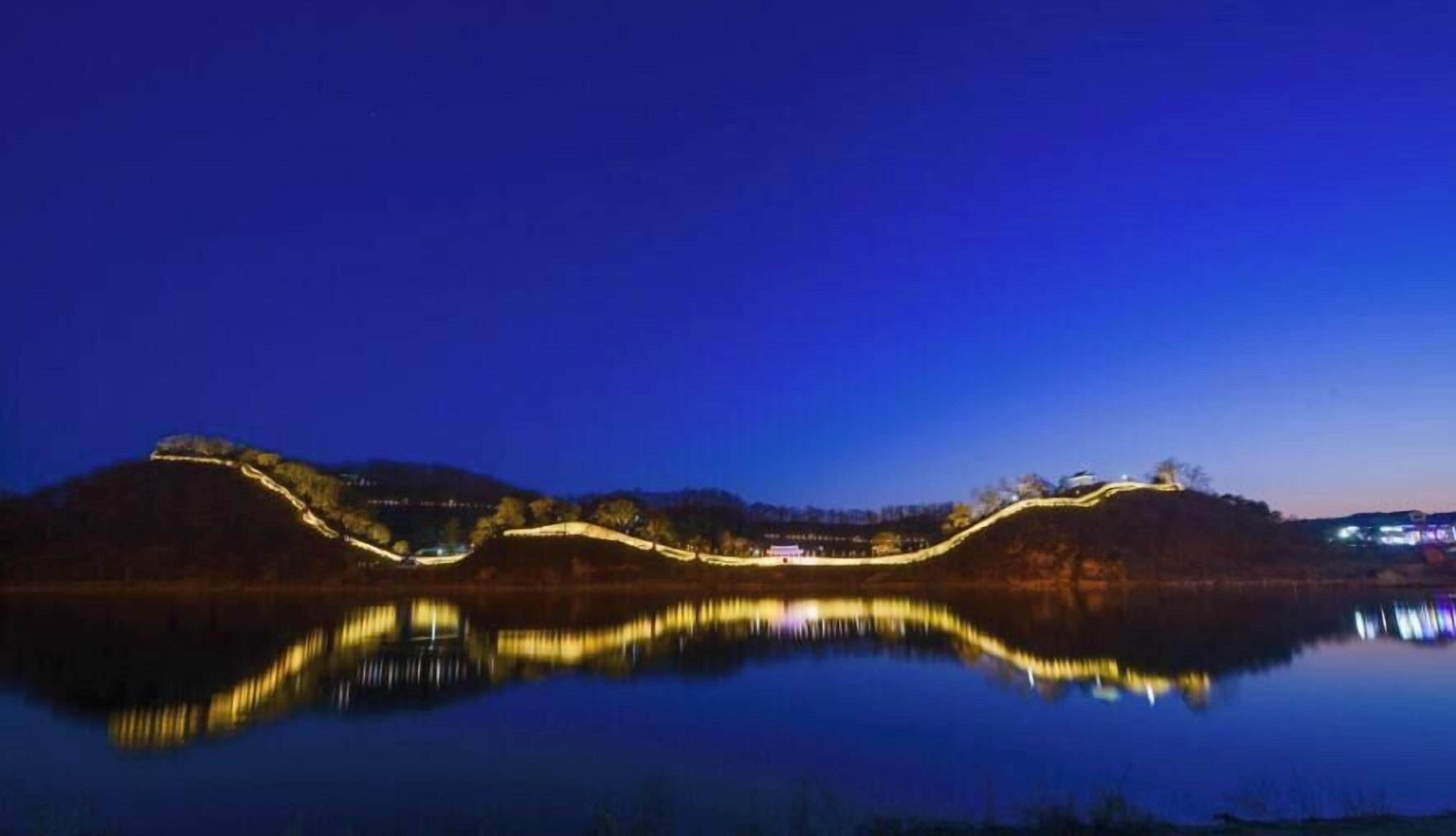 A nighttime view of a calm lake with illuminated structures on the hills.