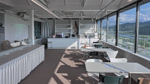 Un espacio de oficina moderno con una zona de bar, ventanas amplias con vistas a un río y montañas, y una variedad de arreglos de asientos.