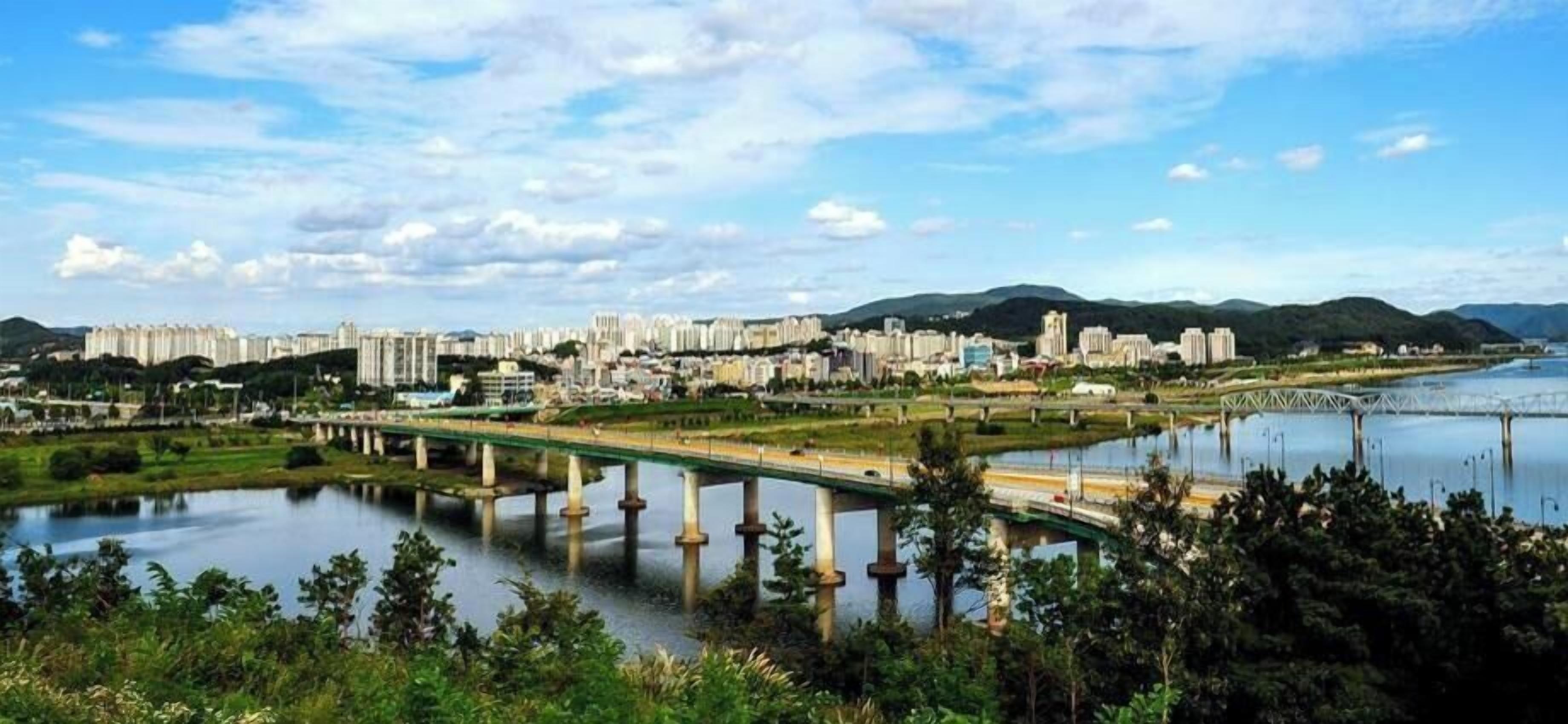 A cityscape with a bridge over a river, surrounded by greenery and hills.