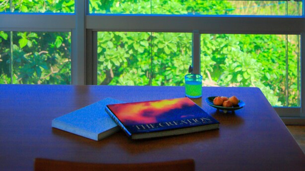 Une table avec un livre et un verre d’eau, un luminaire suspendu et une vue sur l’océan par de grandes fenêtres.