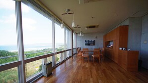 Une pièce spacieuse dotée de grandes fenêtres offrant une vue sur la mer, d’un plancher en bois, d’un coin repas avec des chaises et d’un téléviseur fixé au mur.