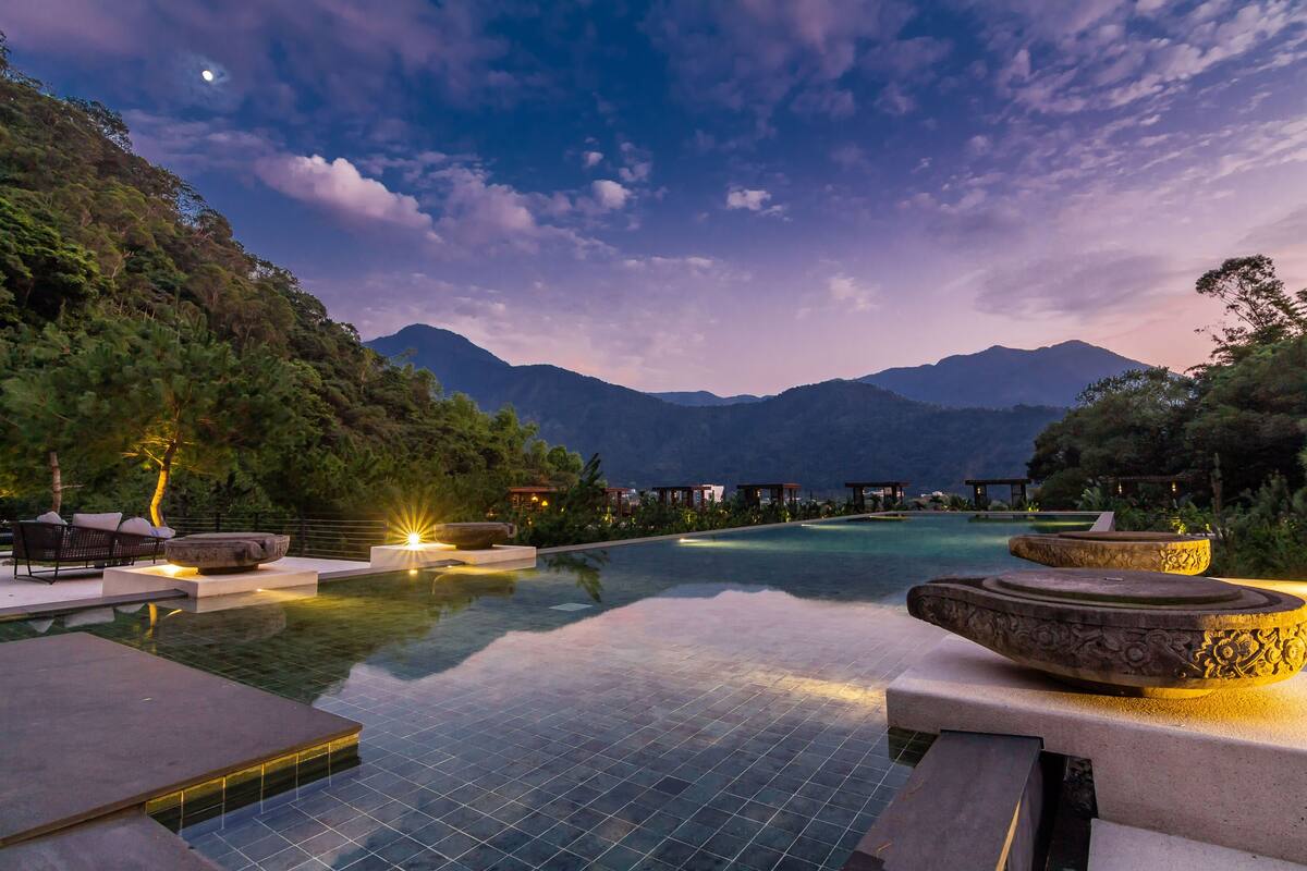 Una piscina infinita tranquila con montañas al fondo, rodeada de exuberante vegetación y mobiliario moderno para exteriores.