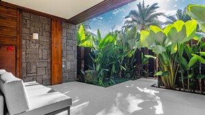 Un espace extérieur moderne aménagé avec des sièges, un mur en pierre et un lambris en bois, entouré d’une végétation tropicale luxuriante et offrant une vue dégagée sur les palmiers.