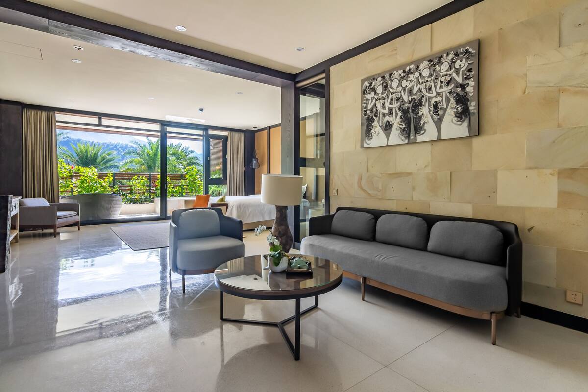 Una sala de estar moderna con un sofá, sillones y una mesa de centro. Hay un ventanal con vista a zonas verdes y un cuadro en la pared.