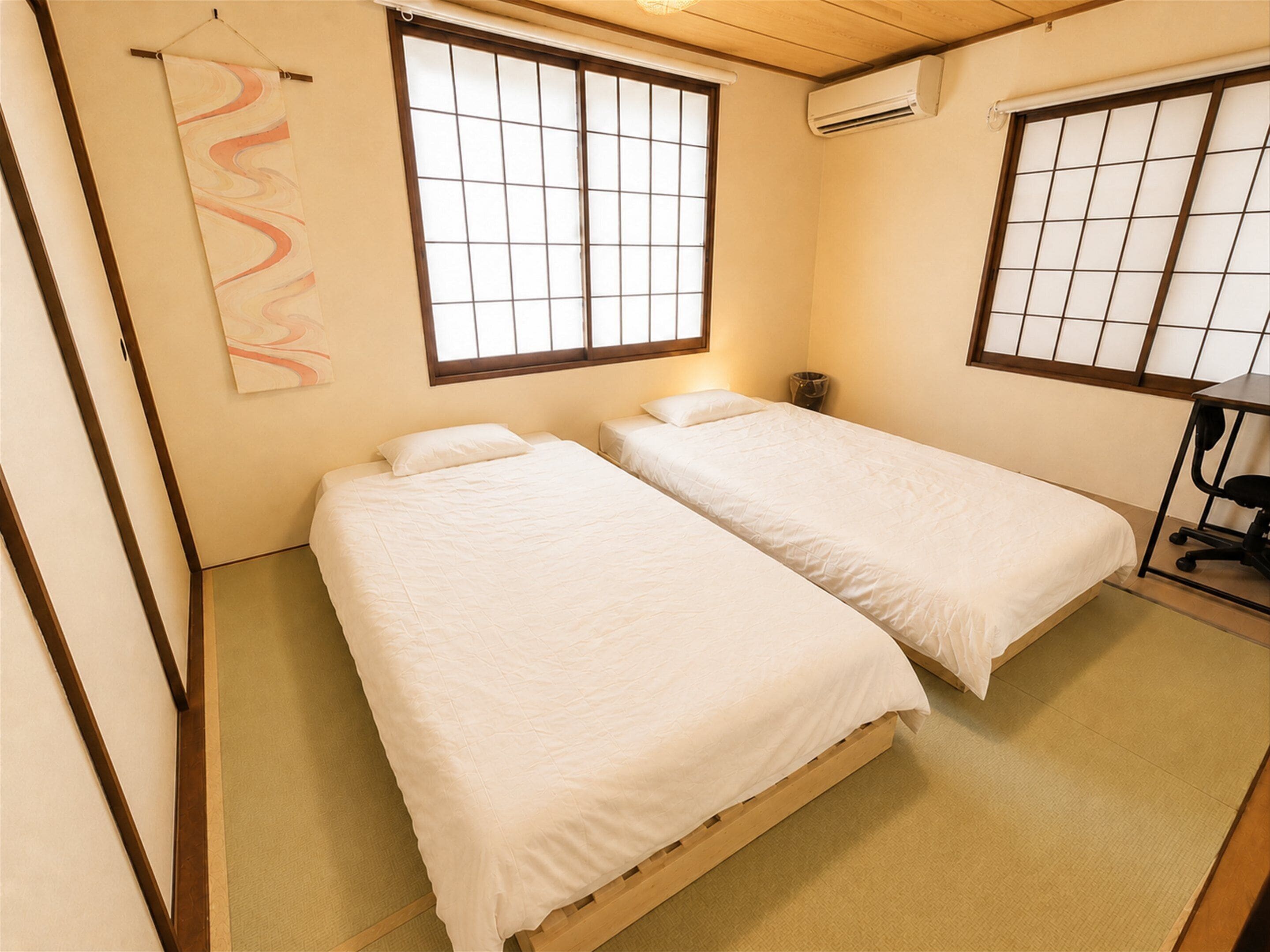 A room with two beds, a window with a grid pattern, an air conditioning unit, and a small desk with a chair.