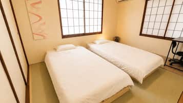 A room with two beds, a window with a grid pattern, an air conditioning unit, and a small desk with a chair.