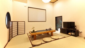 Ein traditioneller japanischer Raum mit Tatami-Bodenbelag, einem niedrigen Tisch mit Kissen, einem Fernseher und einer Schiebetür.