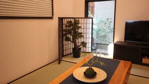 Ein traditioneller japanischer Raum mit Tatami-Boden, einem niedrigen Tisch mit Bonsai, einem Fernseher und einer Schiebetür zu einem Balkon mit Bambuspflanzen.