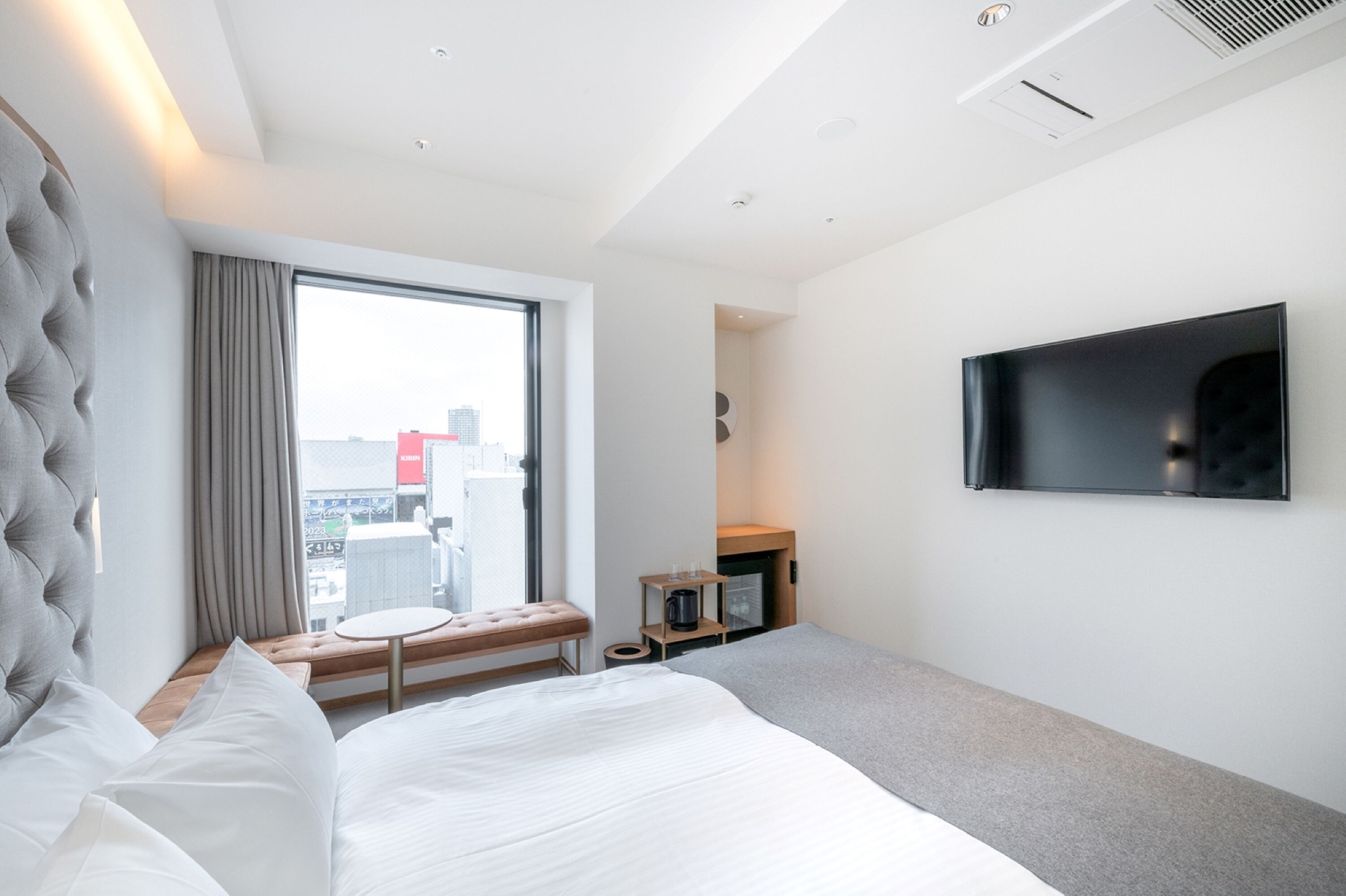 A modern hotel room with a large bed, a flat-screen TV mounted on the wall, a small table, and a window with a view of the city.