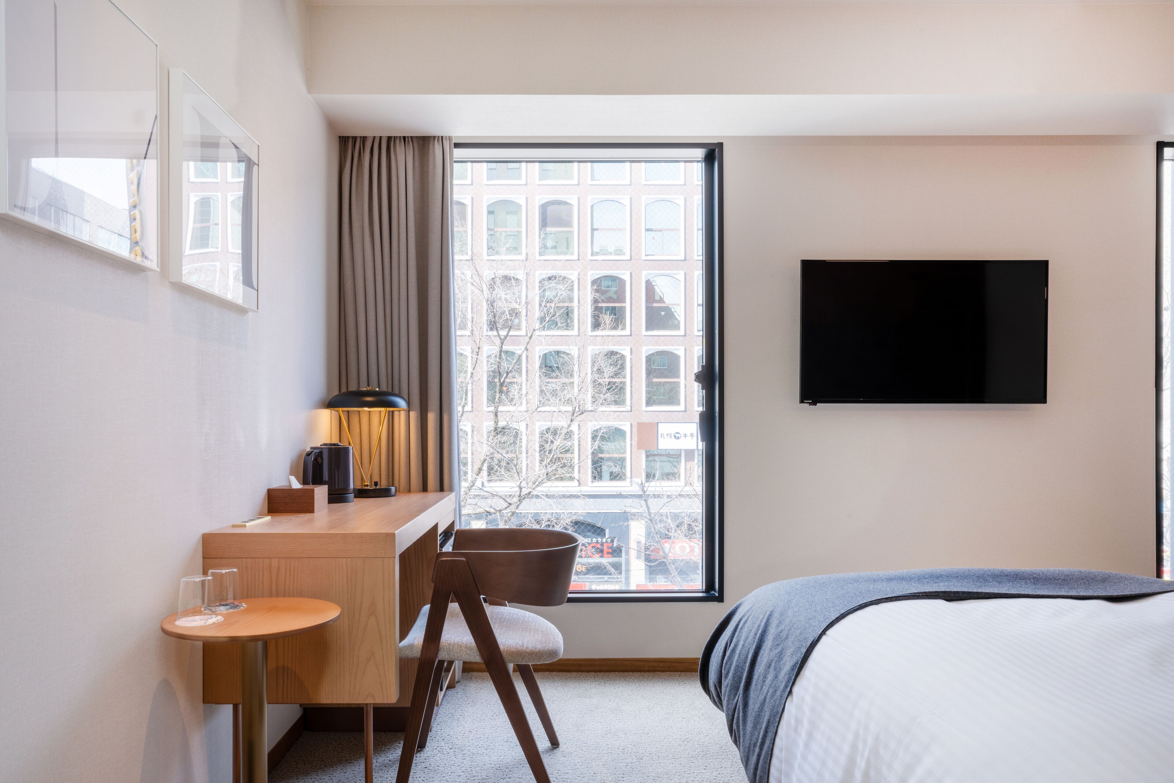 A hotel room with a bed, a desk with a chair, a TV mounted on the wall, and a large window offering a city view.