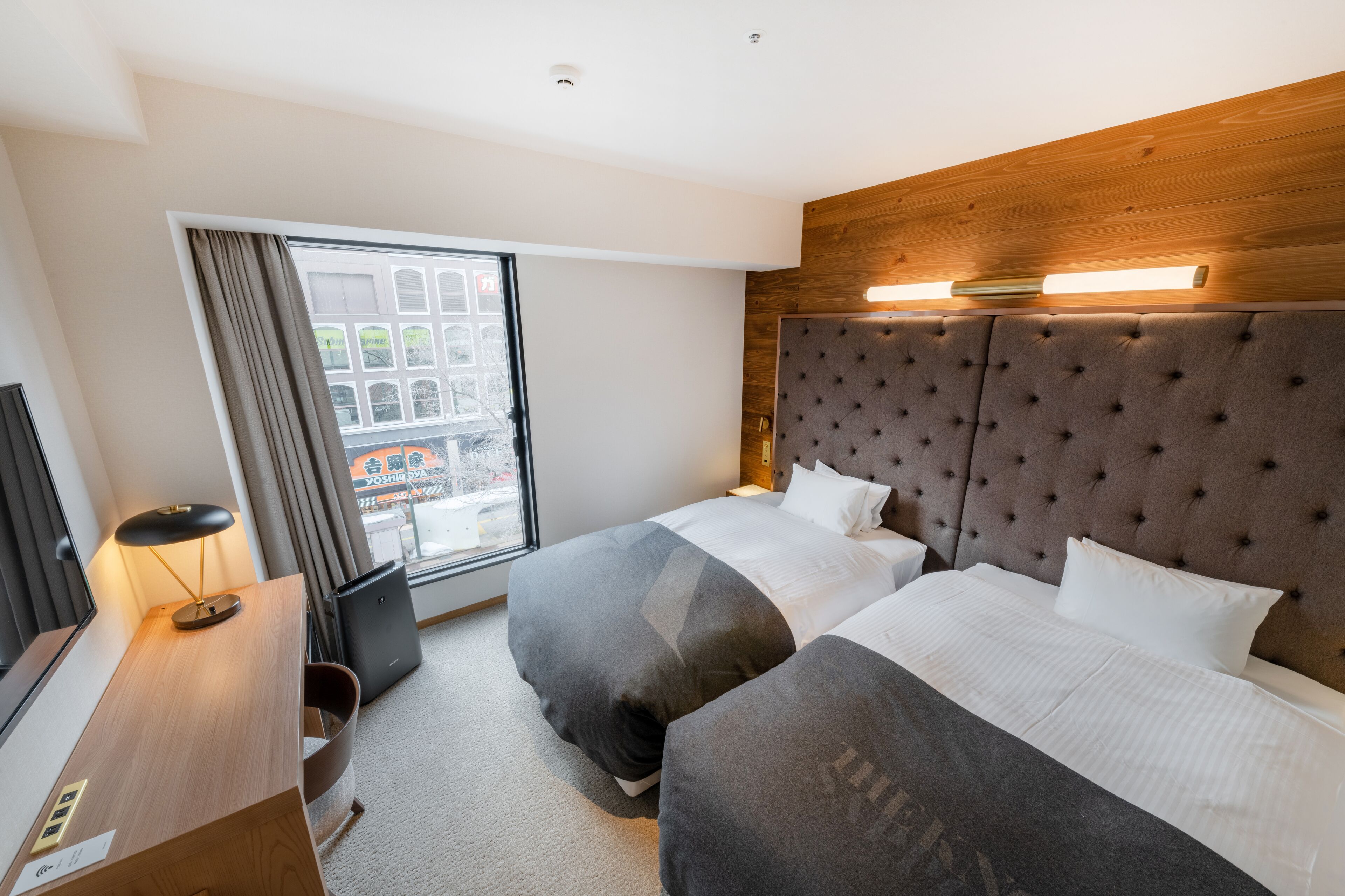A hotel room with two beds, a wooden headboard, a desk with a lamp, and a view of the city through a large window.