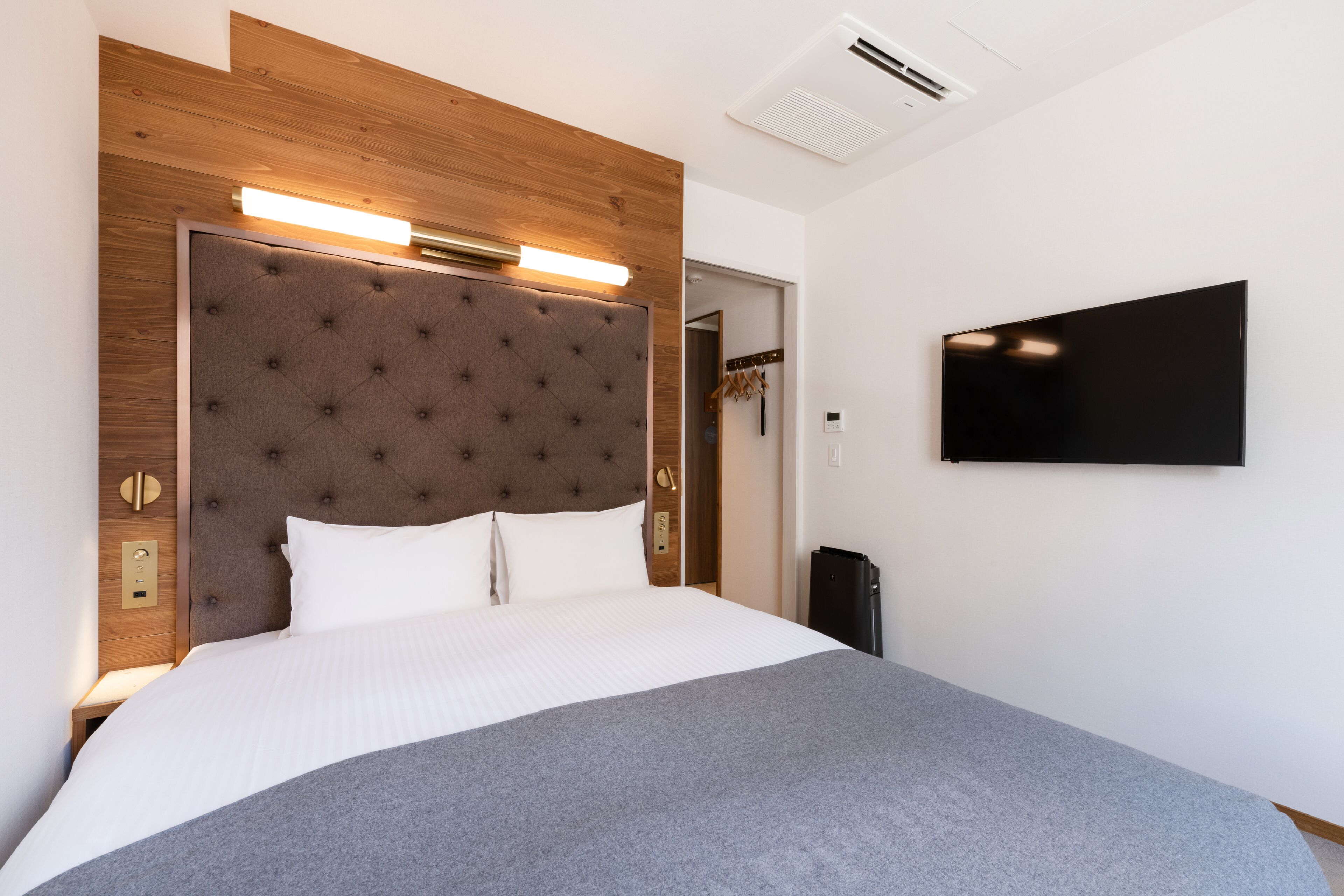 A hotel room with a large bed, a flat-screen TV mounted on the wall, and a wooden headboard with integrated lighting.