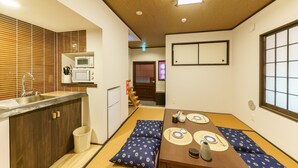 Una tradizionale stanza giapponese con pavimento in tatami, un tavolino in legno basso con cuscini, un angolo cottura con lavandino e microonde, e una porta scorrevole che dà su un'altra stanza.