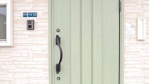 Una puerta verde con un picaporte negro, una pared de piedra, un cartel azul con números y un pequeño dispositivo en la pared.