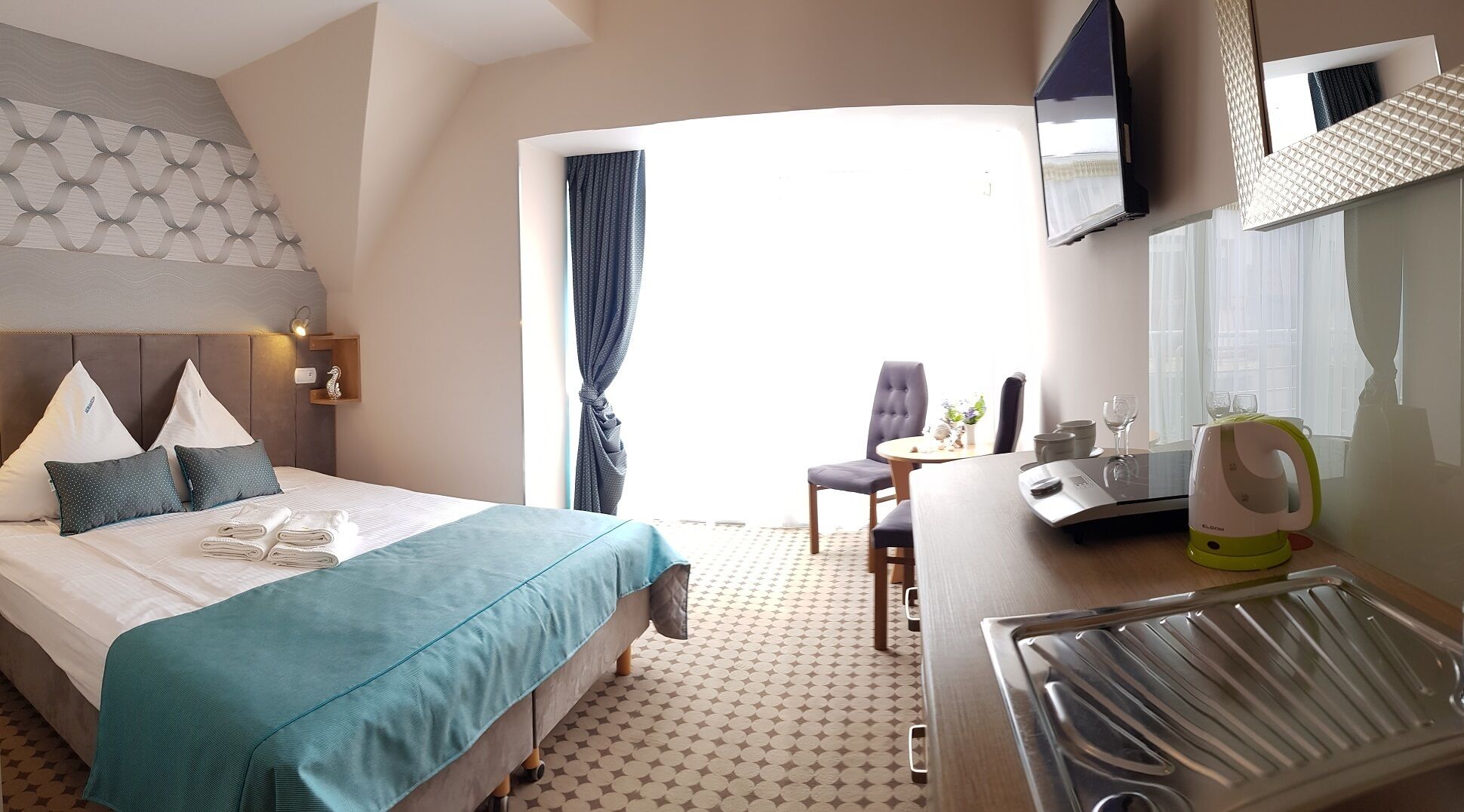 A hotel room with a bed, a desk, a chair, a kettle, and a television.