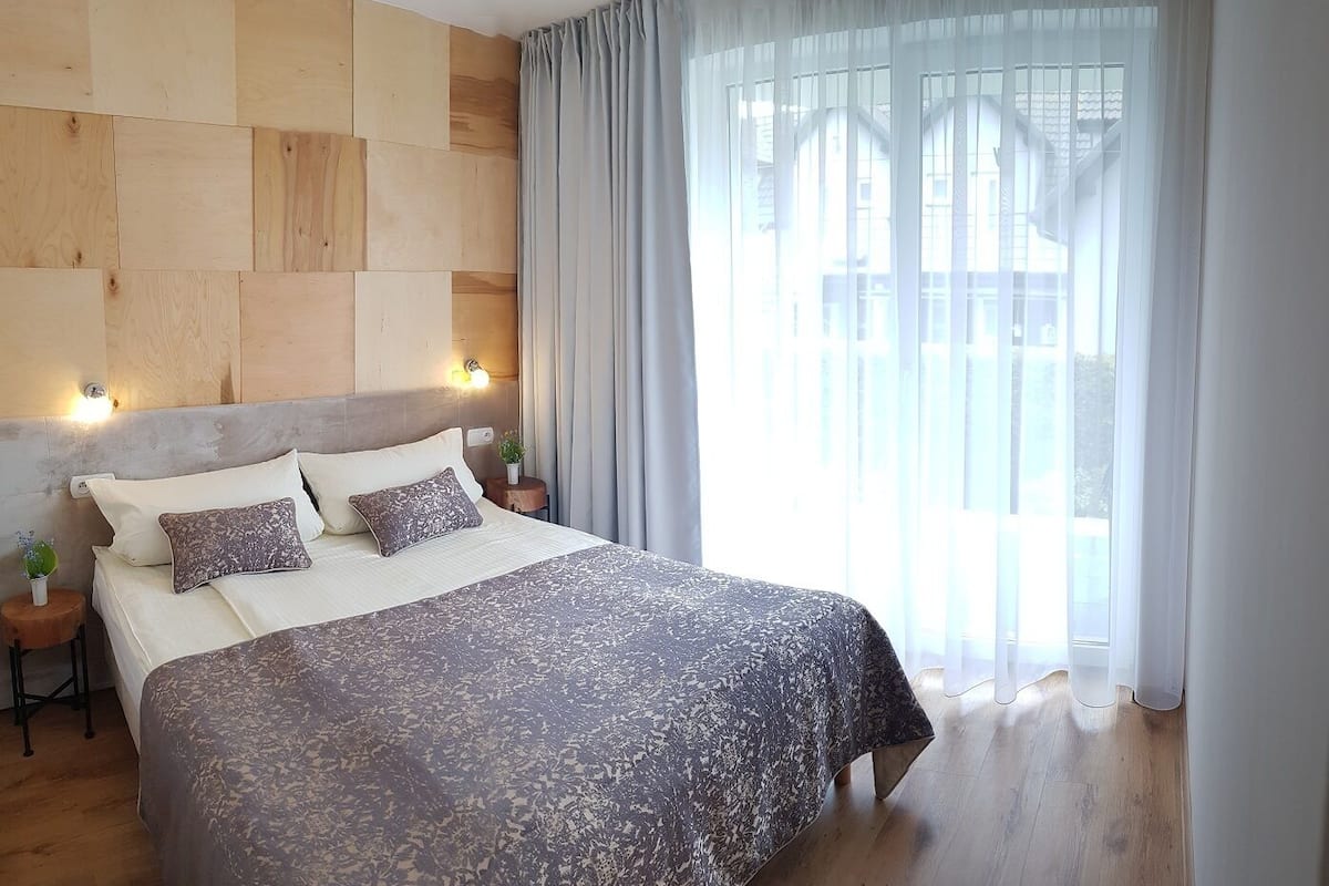 A modern hotel room with a large bed, wooden headboard, two bedside tables with lamps, and a large window with sheer curtains.