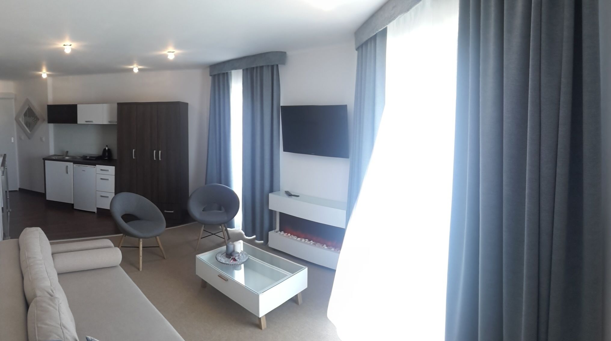 A modern living room with a sofa, two chairs, a coffee table, a TV mounted on the wall, and a kitchen area with cabinets and a sink.