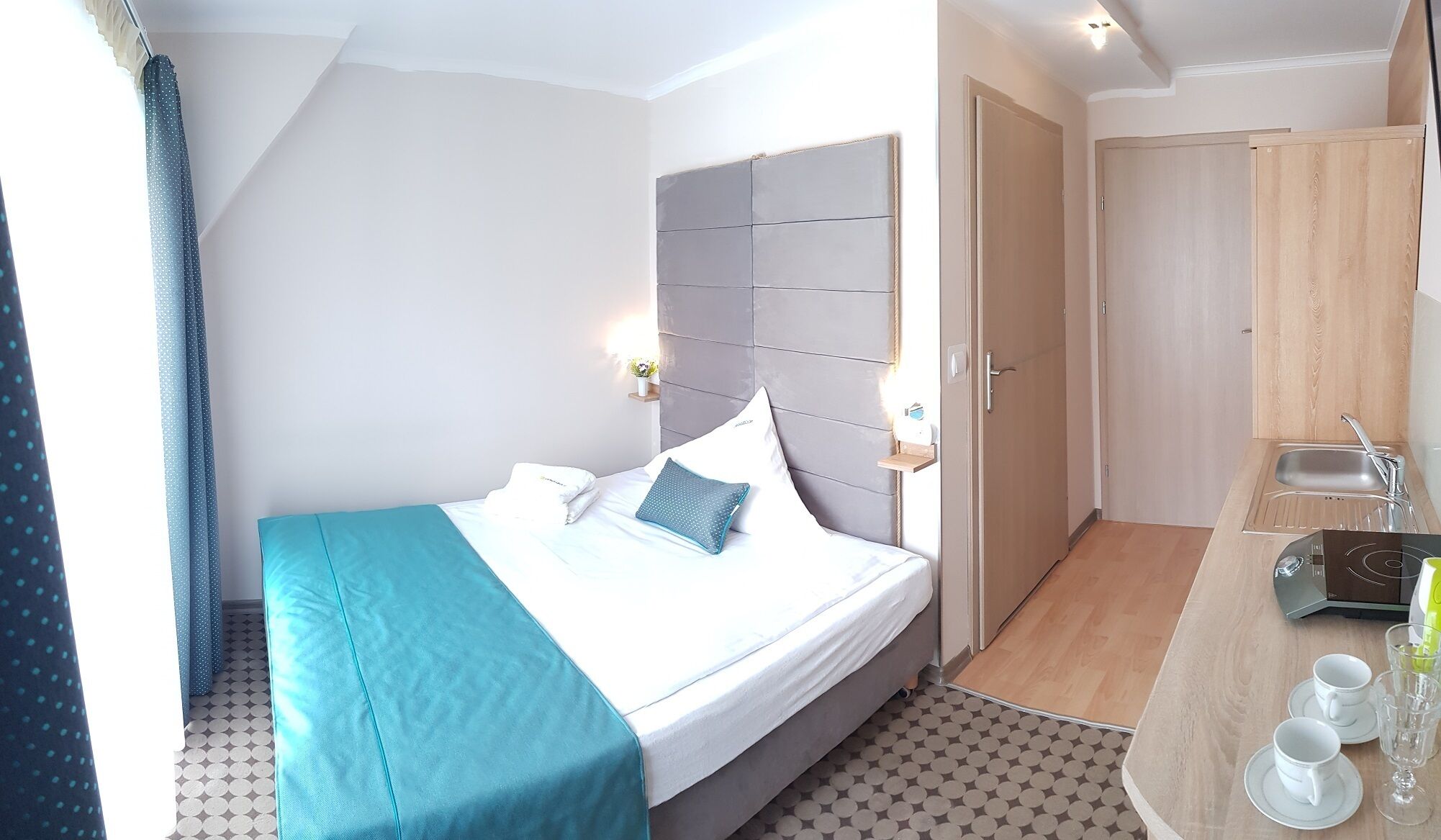 A hotel room with a bed, a bedside table with a lamp, a small shelf with flowers, and a wooden door.