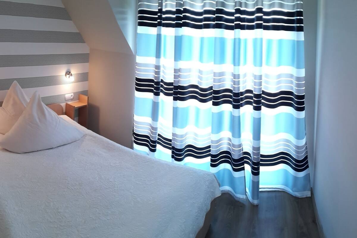 A hotel room with a bed, a nightstand, a wall-mounted light, and a window with striped curtains.
