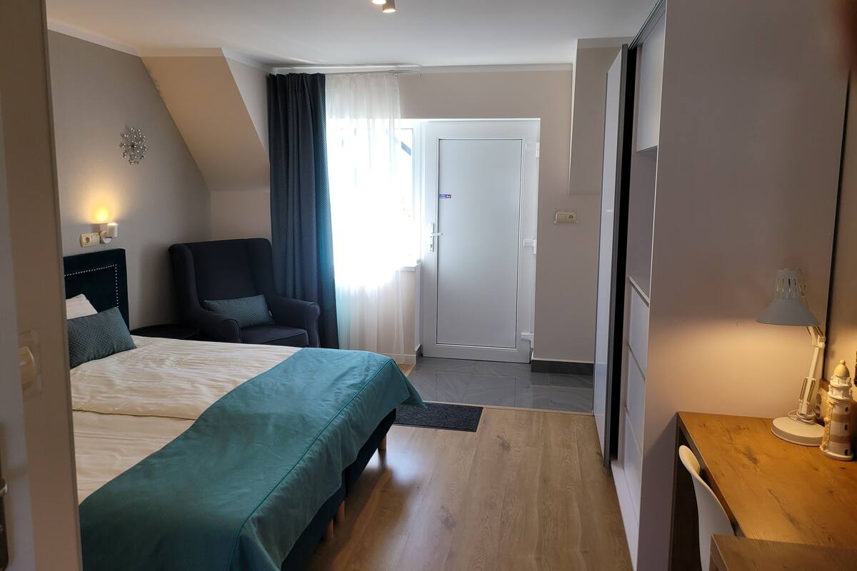 A modern hotel room with a large bed, a desk, a chair, and a door leading to another room.