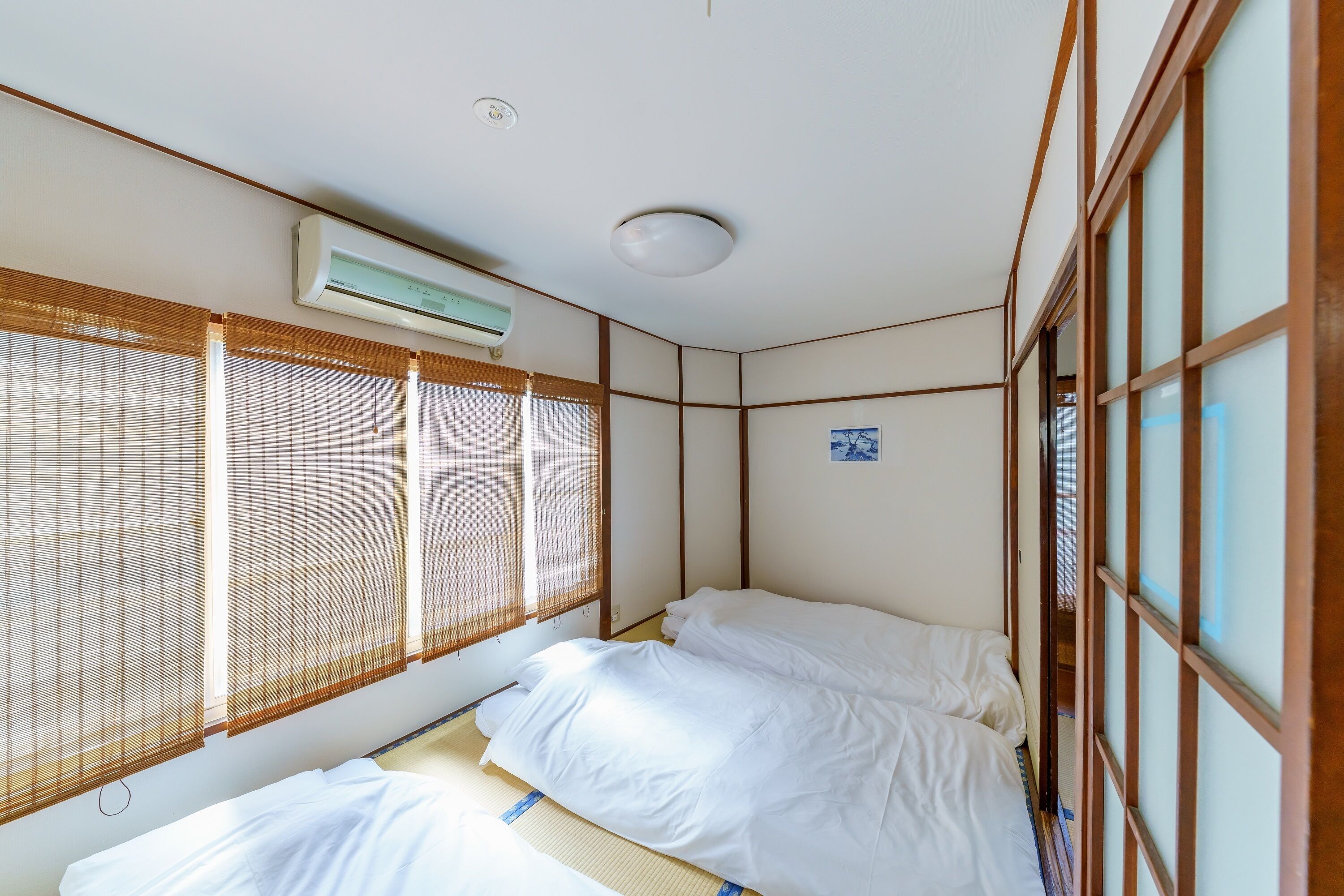 A traditional Japanese-style room with tatami flooring, a futon bed, sliding shoji doors, and a ceiling-mounted air conditioning unit.
