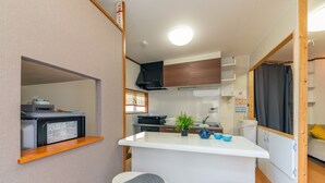 A compact kitchenette with a microwave, a small sink, and a stove. There are two stools with cushions, a potted plant, and a small table with cups. - Yadoya Chigusa Cho (Tokyo)
