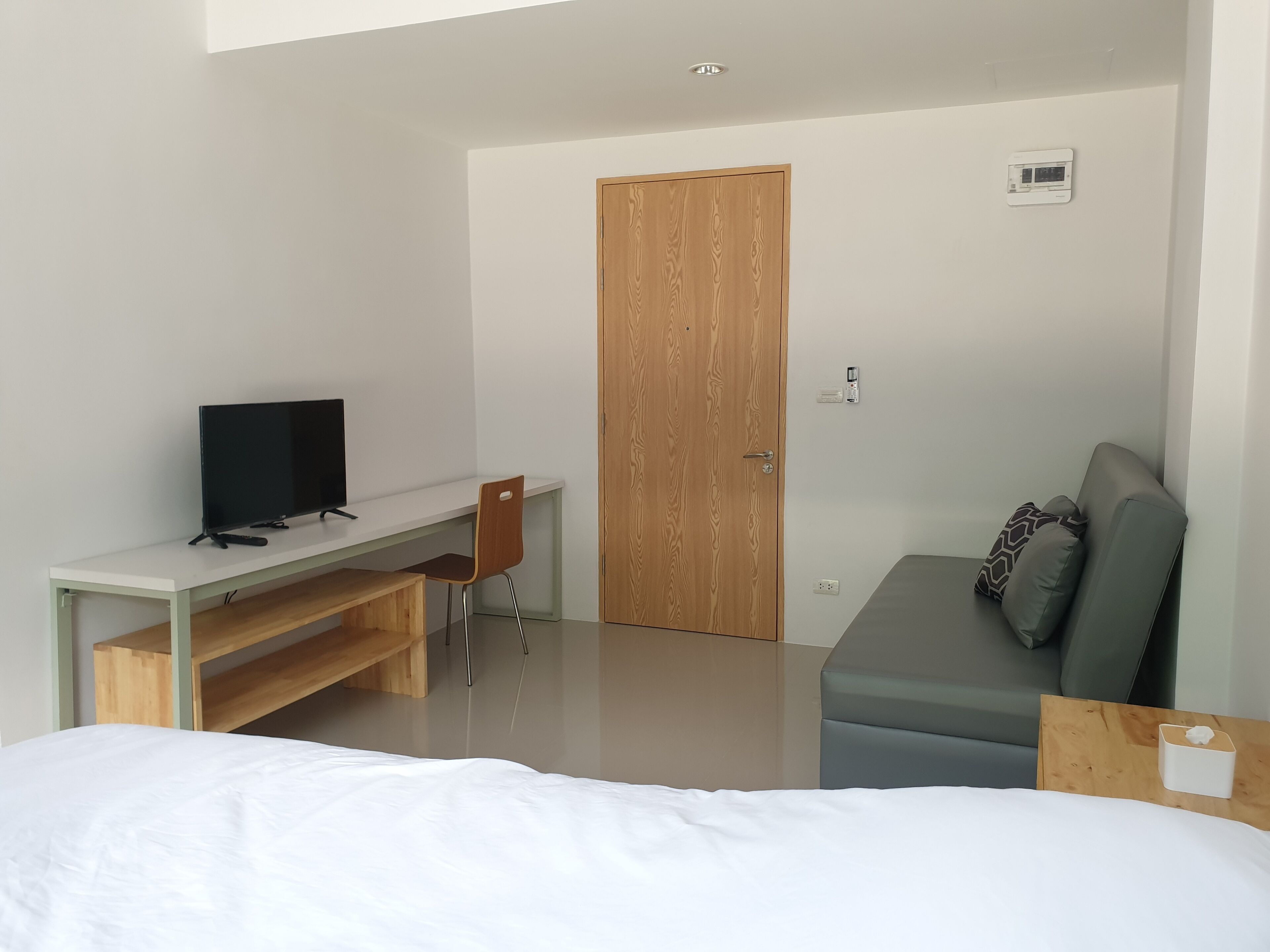 A modern bedroom with a bed, a desk with a TV, a wooden door, and a grey sofa.