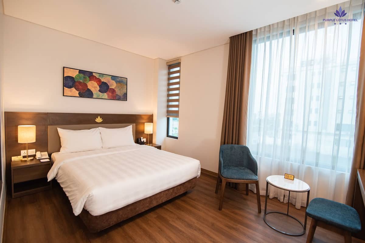 Une chambre d’hôtel avec un grand lit, deux lampes de chevet, une petite table, une chaise et une fenêtre avec des rideaux.
