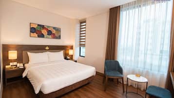 A hotel room with a large bed, two bedside lamps, a small table, a chair, and a window with curtains.