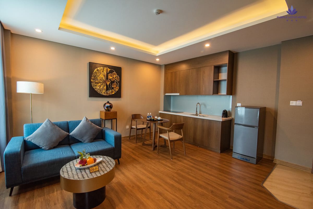 Une chambre d’hôtel moderne avec un canapé bleu, une petite table à manger, un coin cuisine et un réfrigérateur.