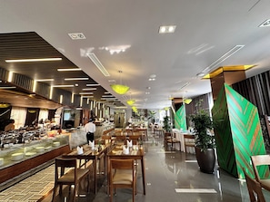A modern hotel lobby with a marble reception desk, multiple dining tables, and a variety of plants.