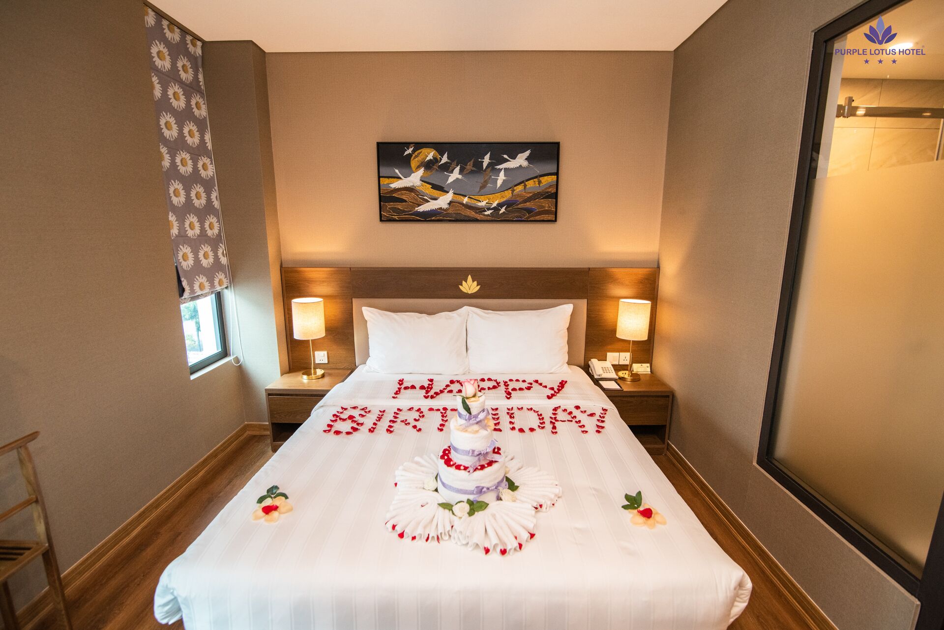 A hotel room with a bed, two bedside tables, a nightstand, a window with curtains, a painting on the wall, and a door.