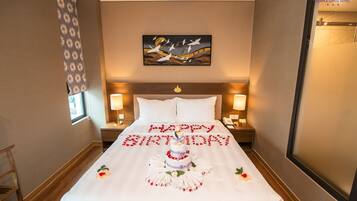 A hotel room with a bed, two bedside tables, a nightstand, a window with curtains, a painting on the wall, and a door.