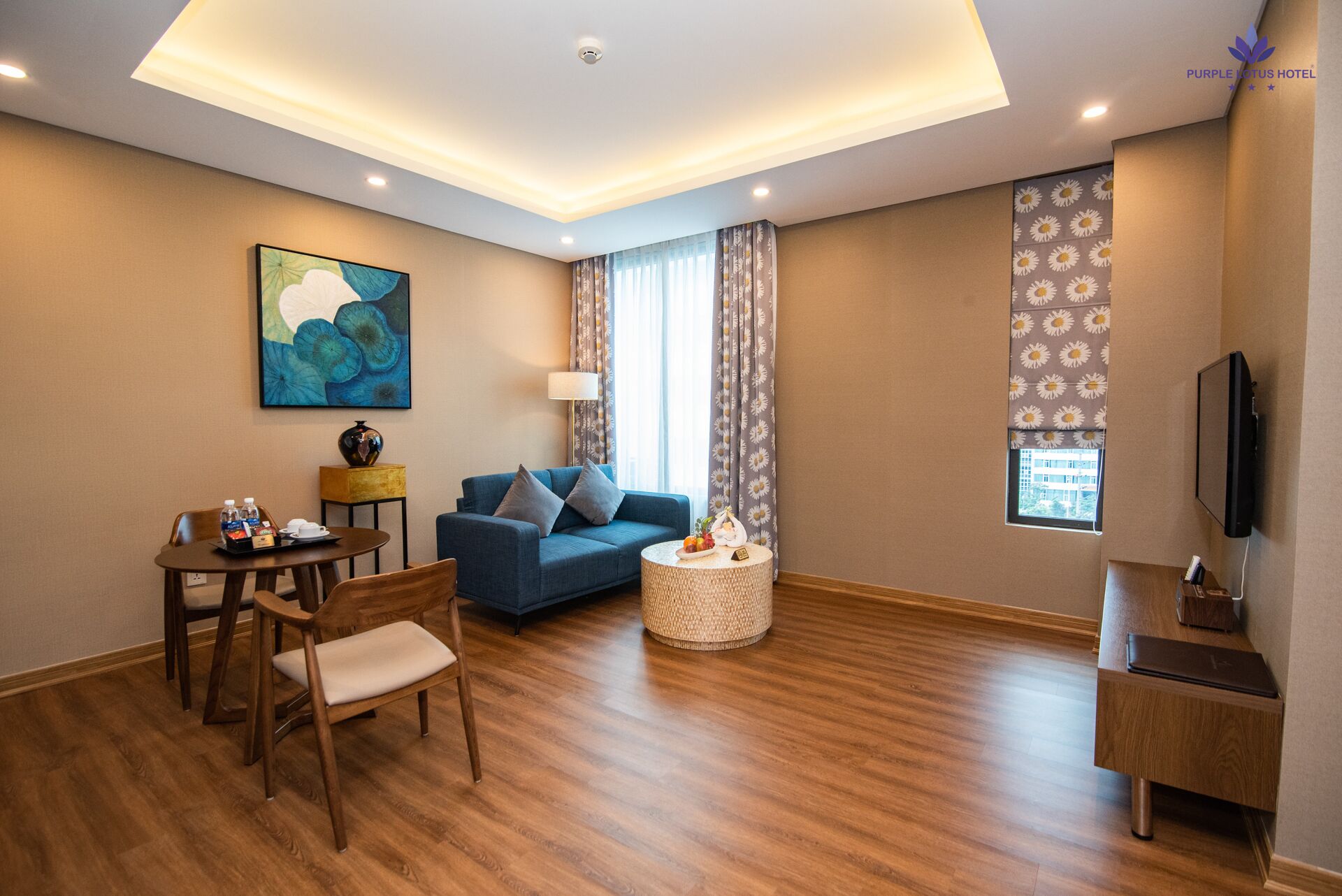 A modern hotel room with a blue sofa, a dining table with chairs, a small round coffee table, and a flat-screen TV.