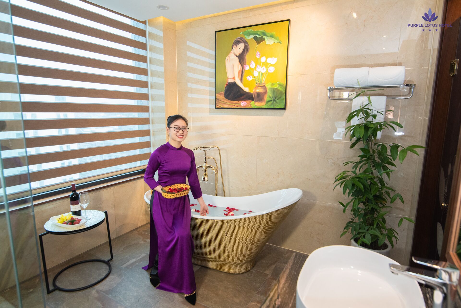 A woman in a purple dress standing in a bathroom with a freestanding bathtub, a sink, and a potted plant.